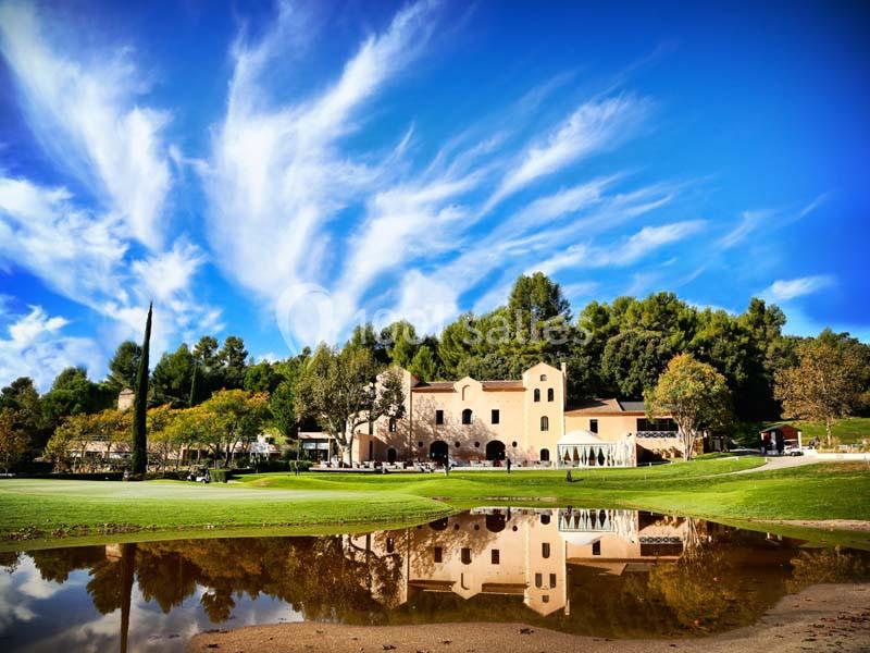 Bâtiment beige entouré de verdure, reflété dans un plan d'eau sous un ciel bleu avec des nuages légers.