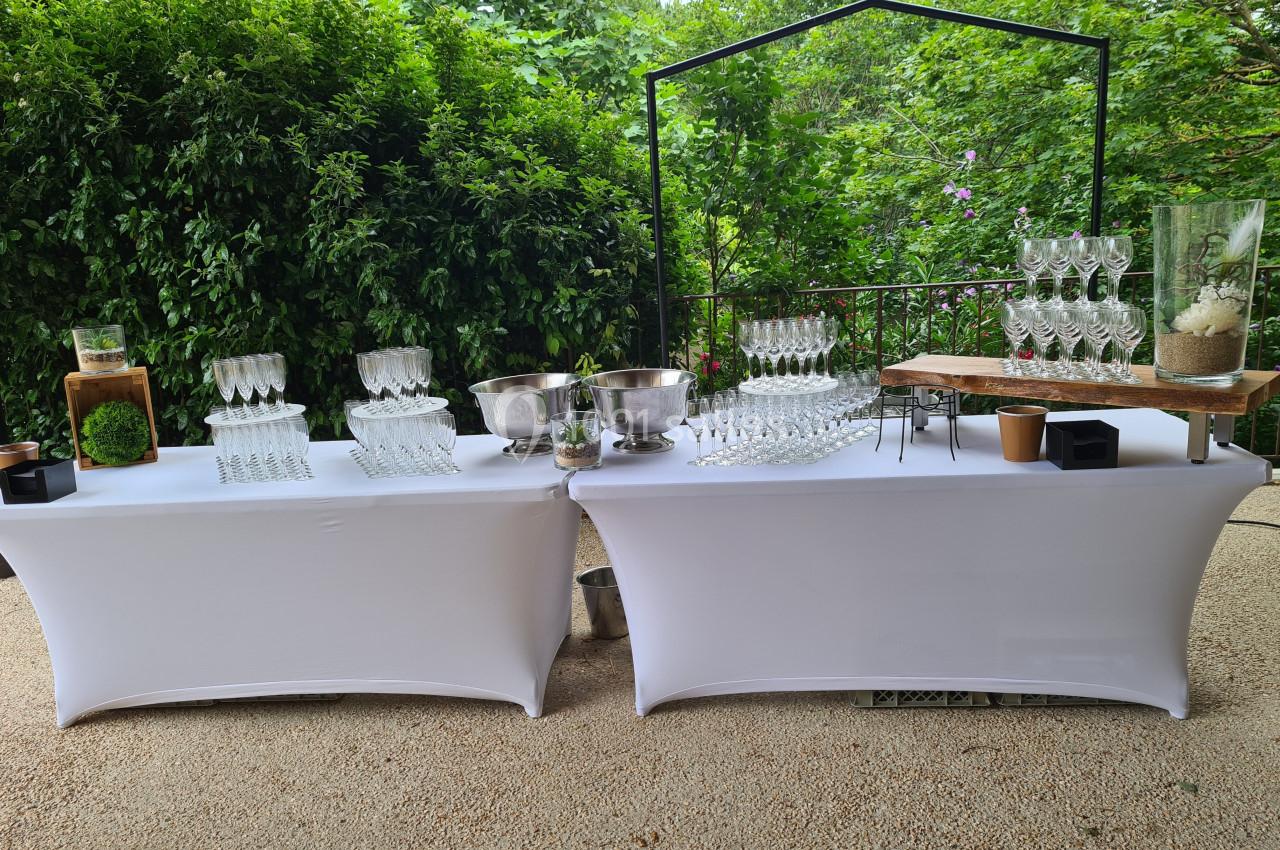 Tables décorées avec nappes blanches, verres, seaux à glace et éléments naturels, disposées en extérieur verdoyant.