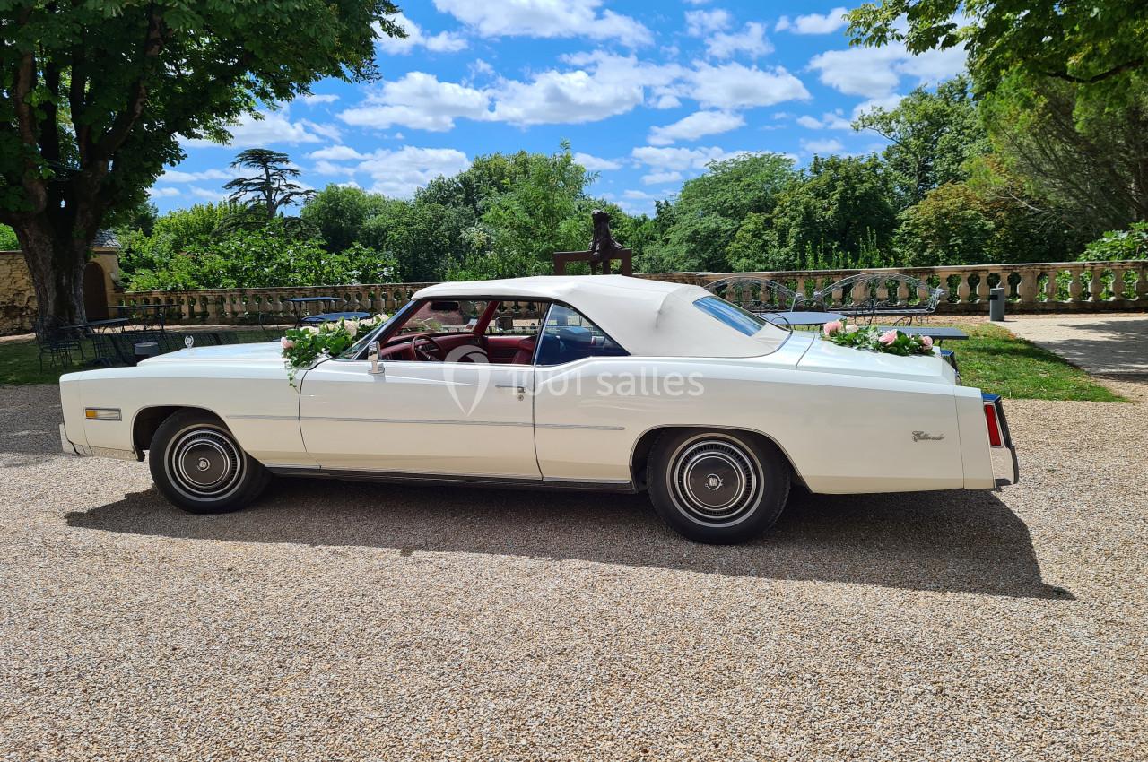Voiture ancienne blanche décorée de fleurs, stationnée sur une allée gravillonnée devant un jardin arboré.