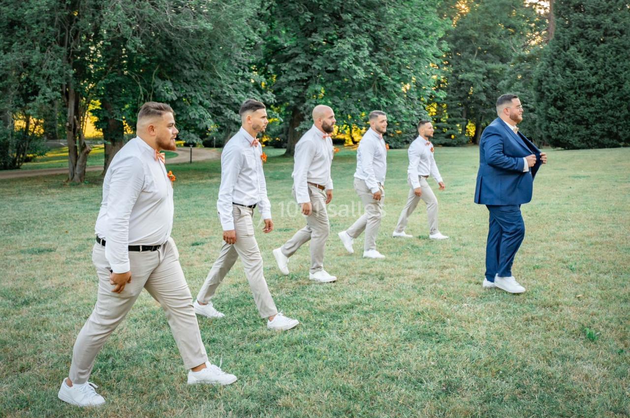 Cinq hommes en chemises blanches et pantalons beiges marchent sur une pelouse derrière un homme en costume bleu.