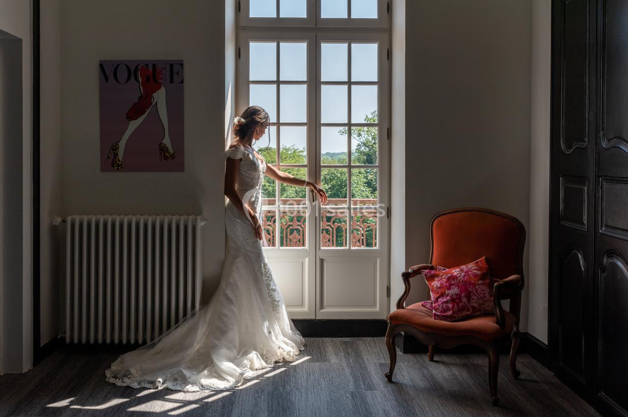 Une femme en robe de mariée regarde par une porte-fenêtre, éclairée par la lumière naturelle, dans une pièce élégante.