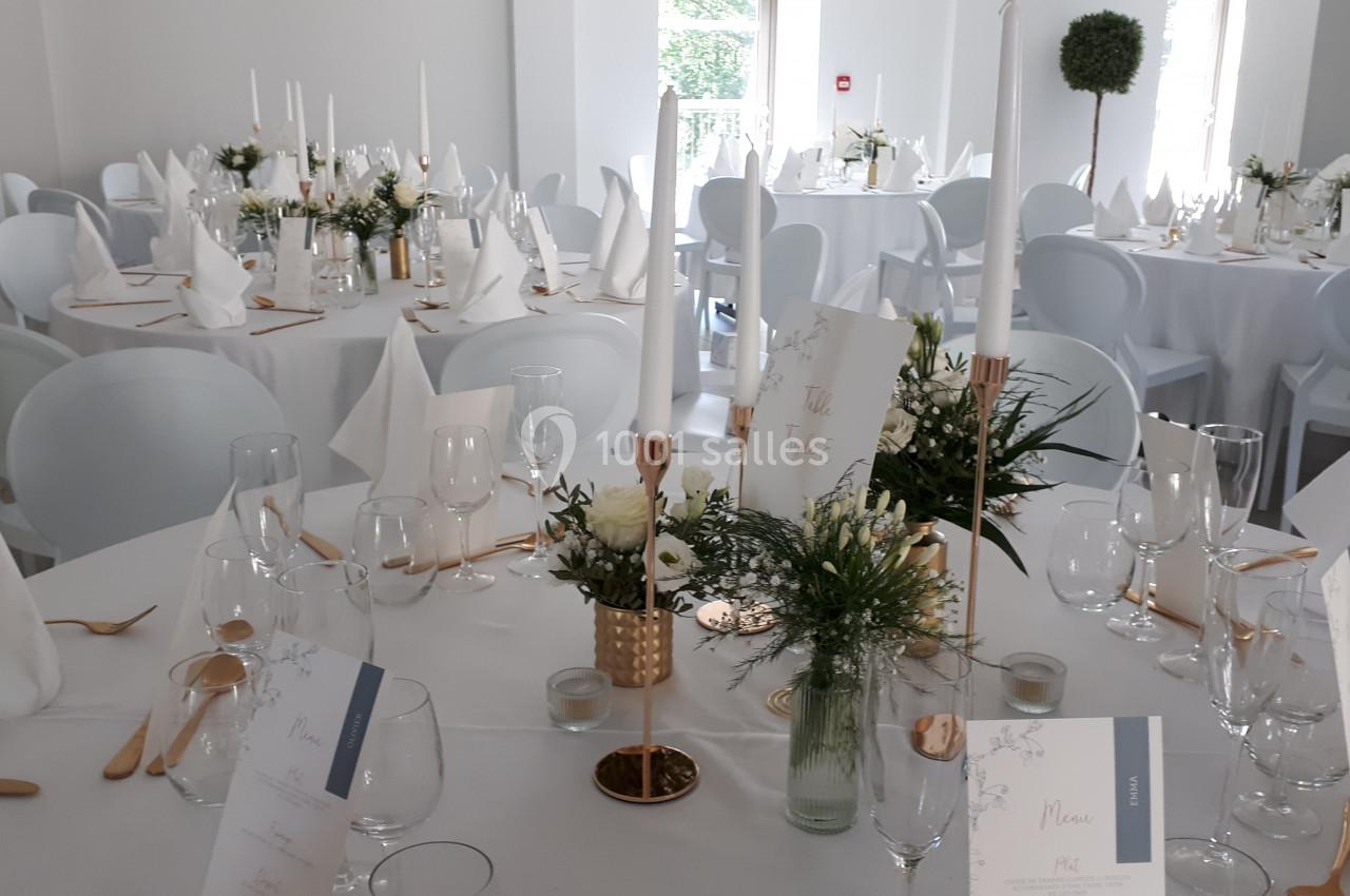 Salle de réception décorée avec des tables rondes, nappes blanches, bougies, fleurs et serviettes pliées.