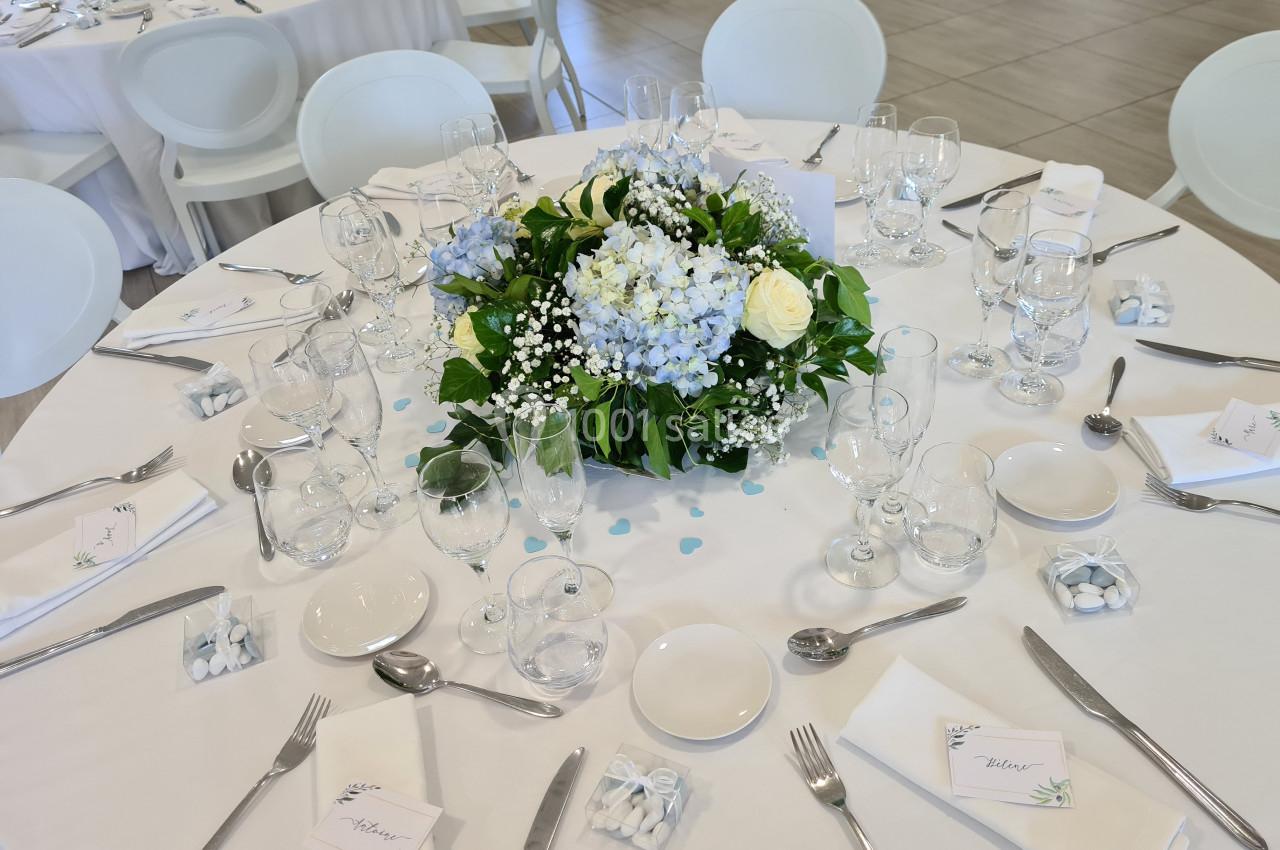 Table ronde dressée avec une nappe blanche, centre de table floral bleu et blanc, couverts et verres disposés.