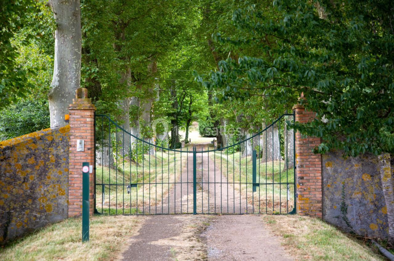Portail en fer forgé ouvert, encadré de piliers en briques, donnant sur une allée bordée d'arbres verdoyants.