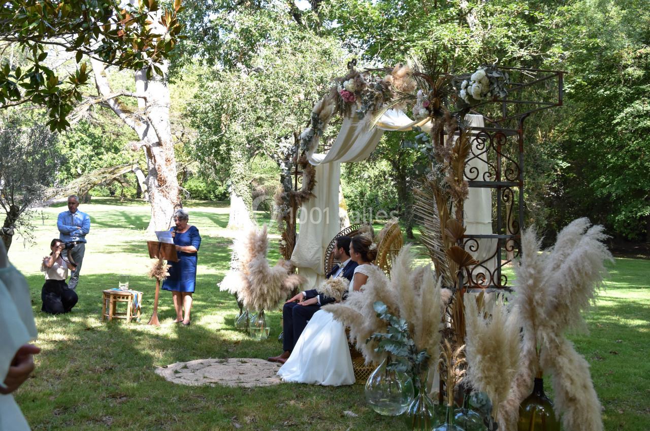Cérémonie de mariage en extérieur avec un couple assis sous une arche décorée de fleurs et de pampas, dans un jardin…