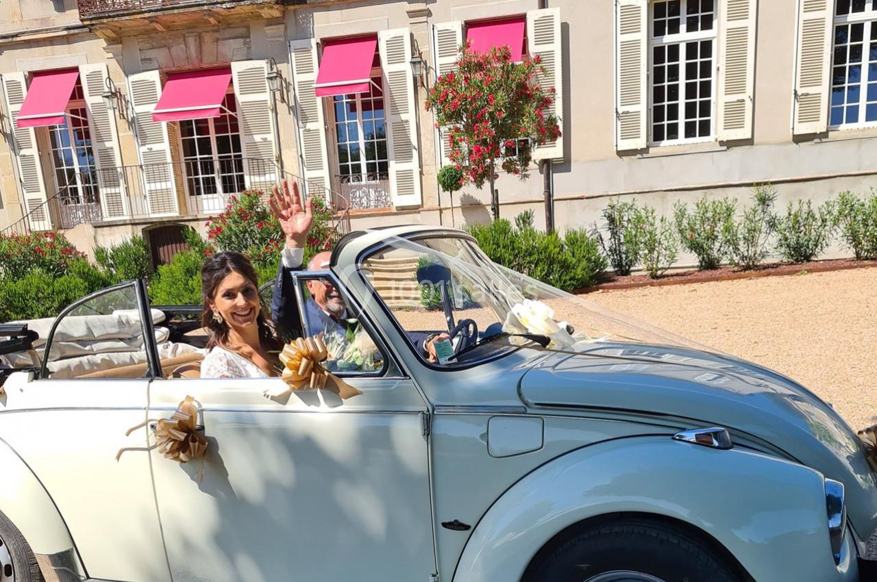 Une mariée souriante salue depuis une voiture décapotable décorée, devant un bâtiment avec volets blancs et stores rouges.