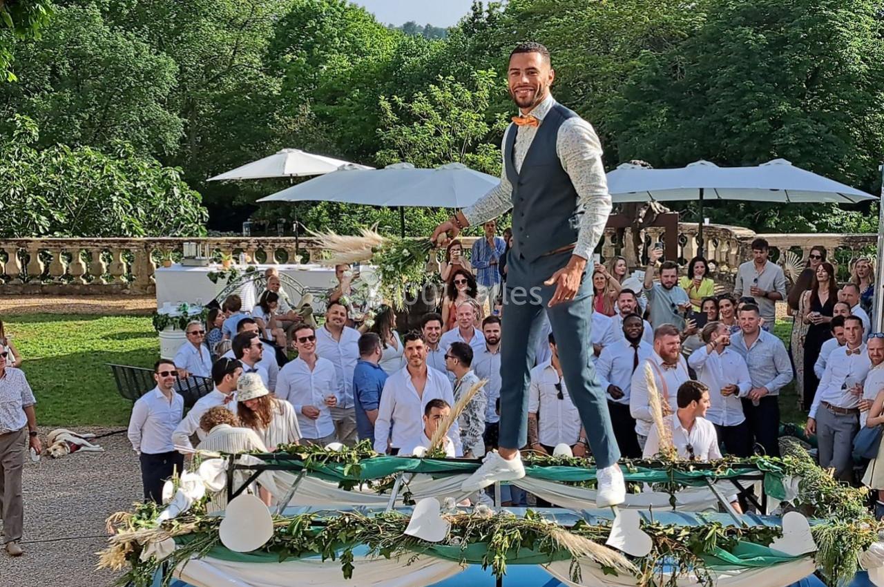 Un homme en costume se tient debout sur une table décorée lors d'un événement en plein air avec des invités en arrière-plan.
