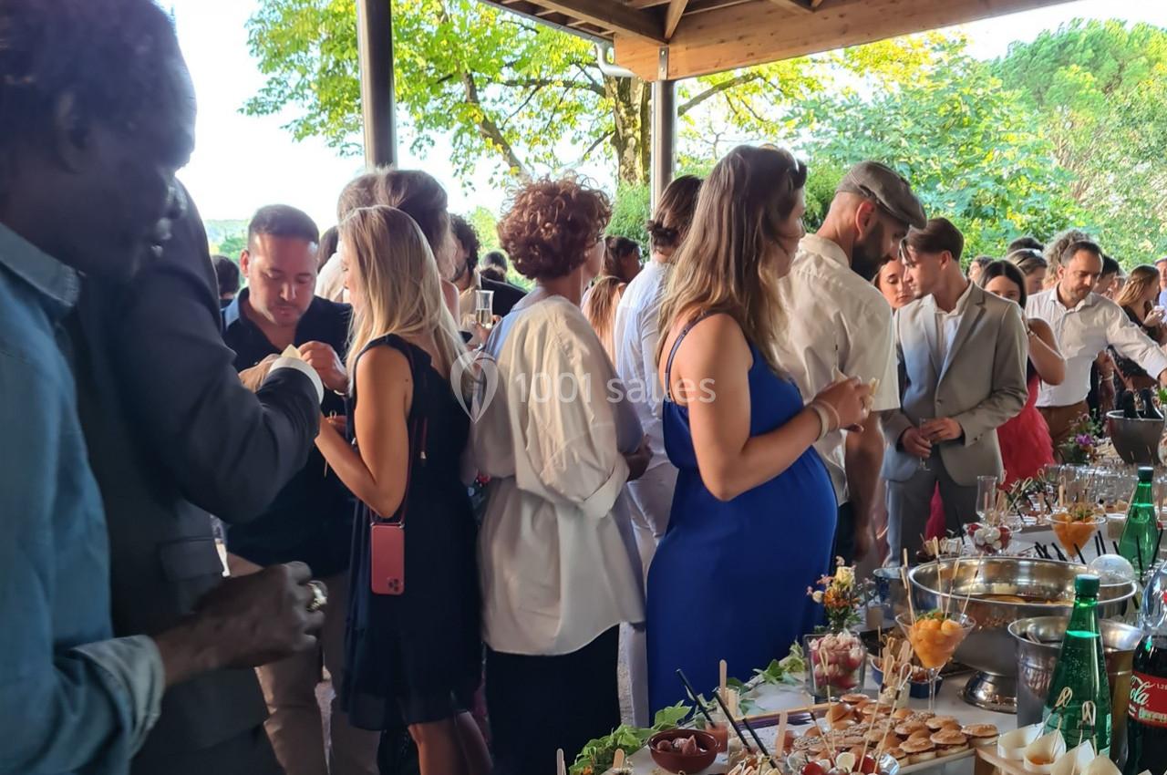 Groupe de personnes discutant autour d'un buffet en extérieur sous un abri, avec des arbres en arrière-plan.