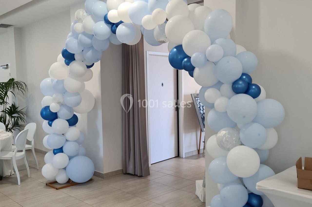 Arche décorative en ballons blancs et bleus installée dans une salle lumineuse avec des murs clairs.