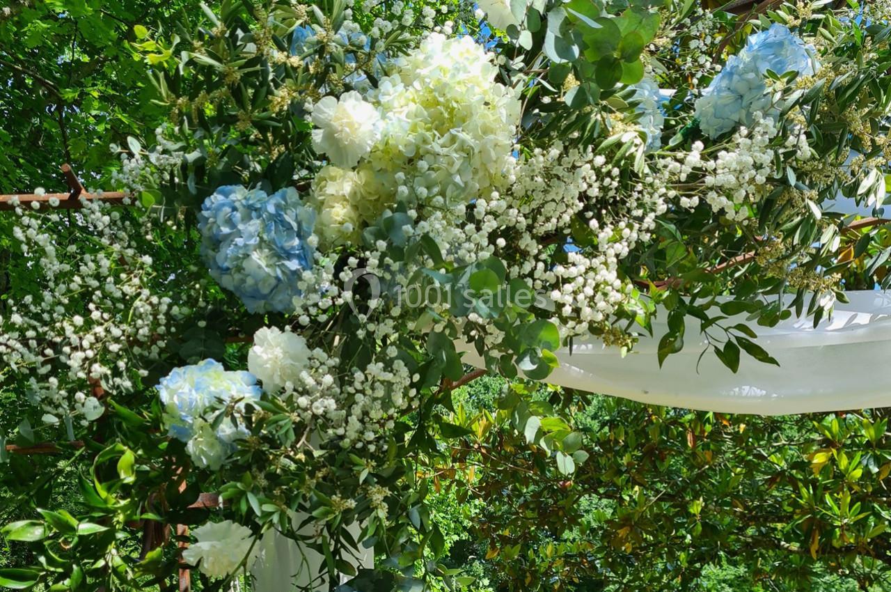 Arrangement floral avec hortensias bleus, gypsophiles et feuillage vert, suspendu sous un voile blanc en extérieur.