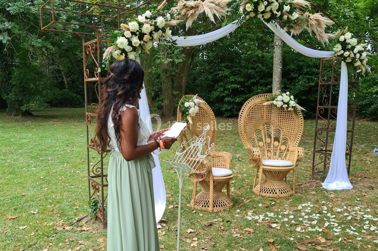Une femme en robe verte se tient devant une arche décorée de fleurs et de chaises en osier dans un jardin.
