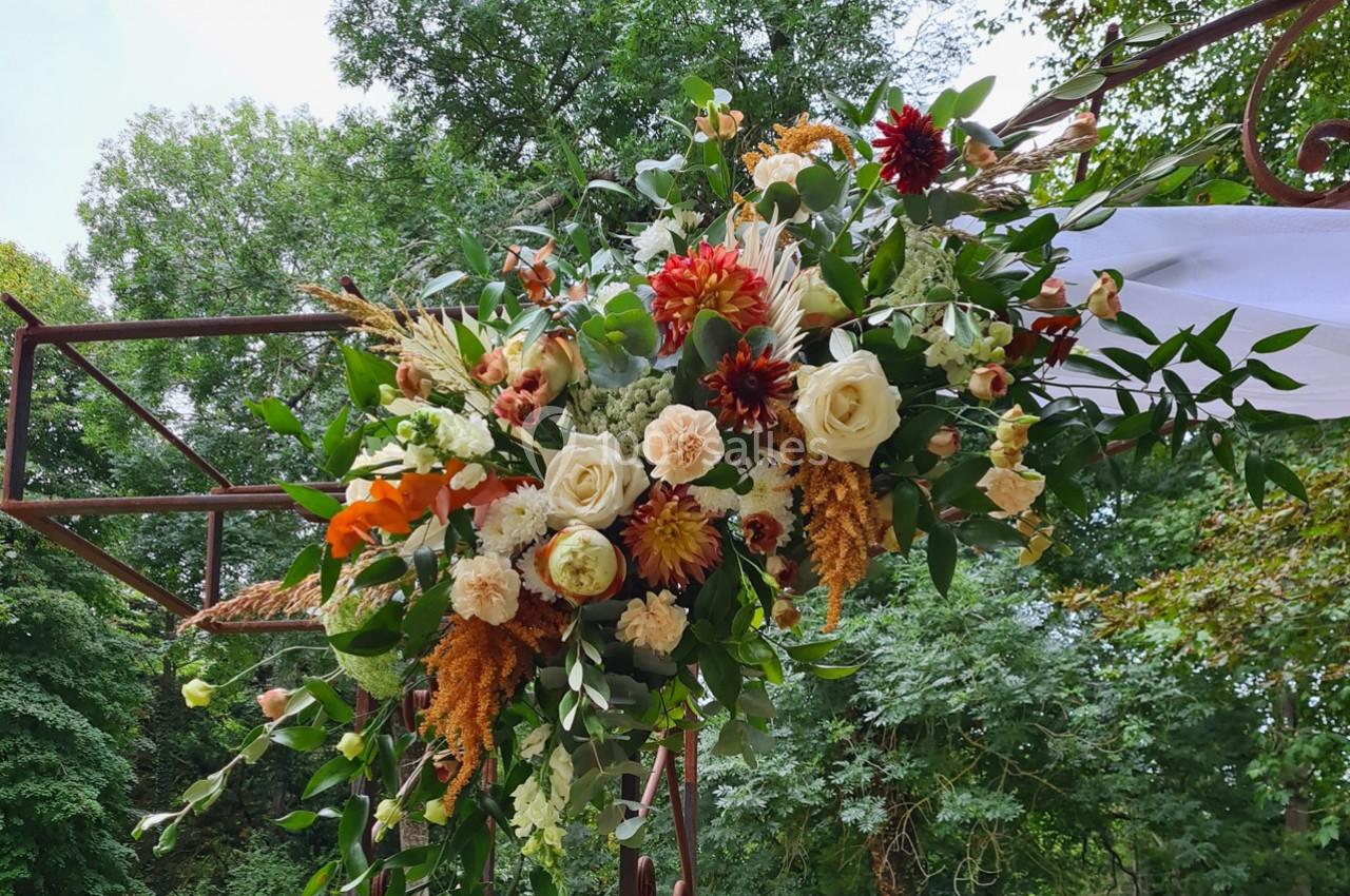 Arrangement floral coloré avec roses, dahlias et feuillage, suspendu à une structure métallique en extérieur verdoyant.