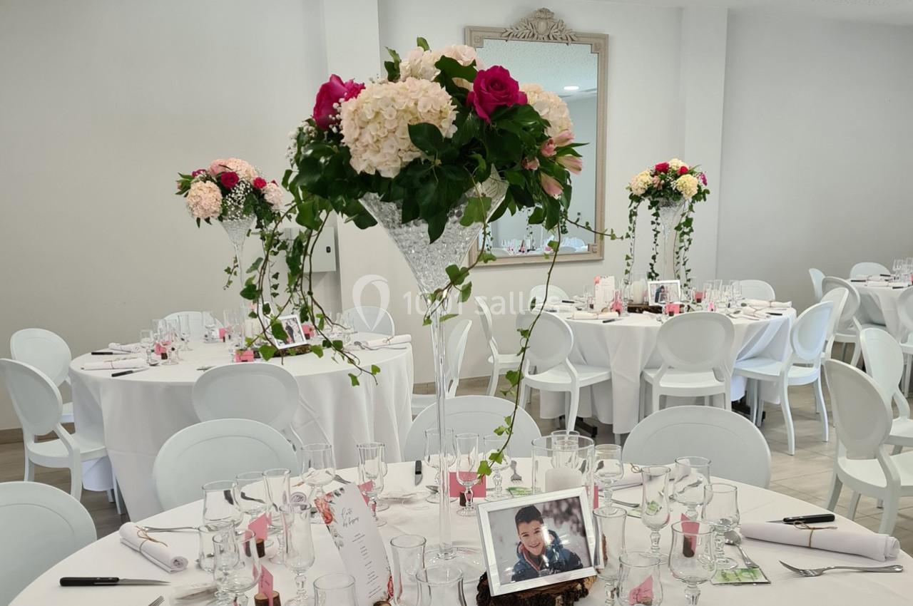 Salle de réception décorée avec des tables rondes, nappes blanches, centres de table fleuris et photo encadrée.