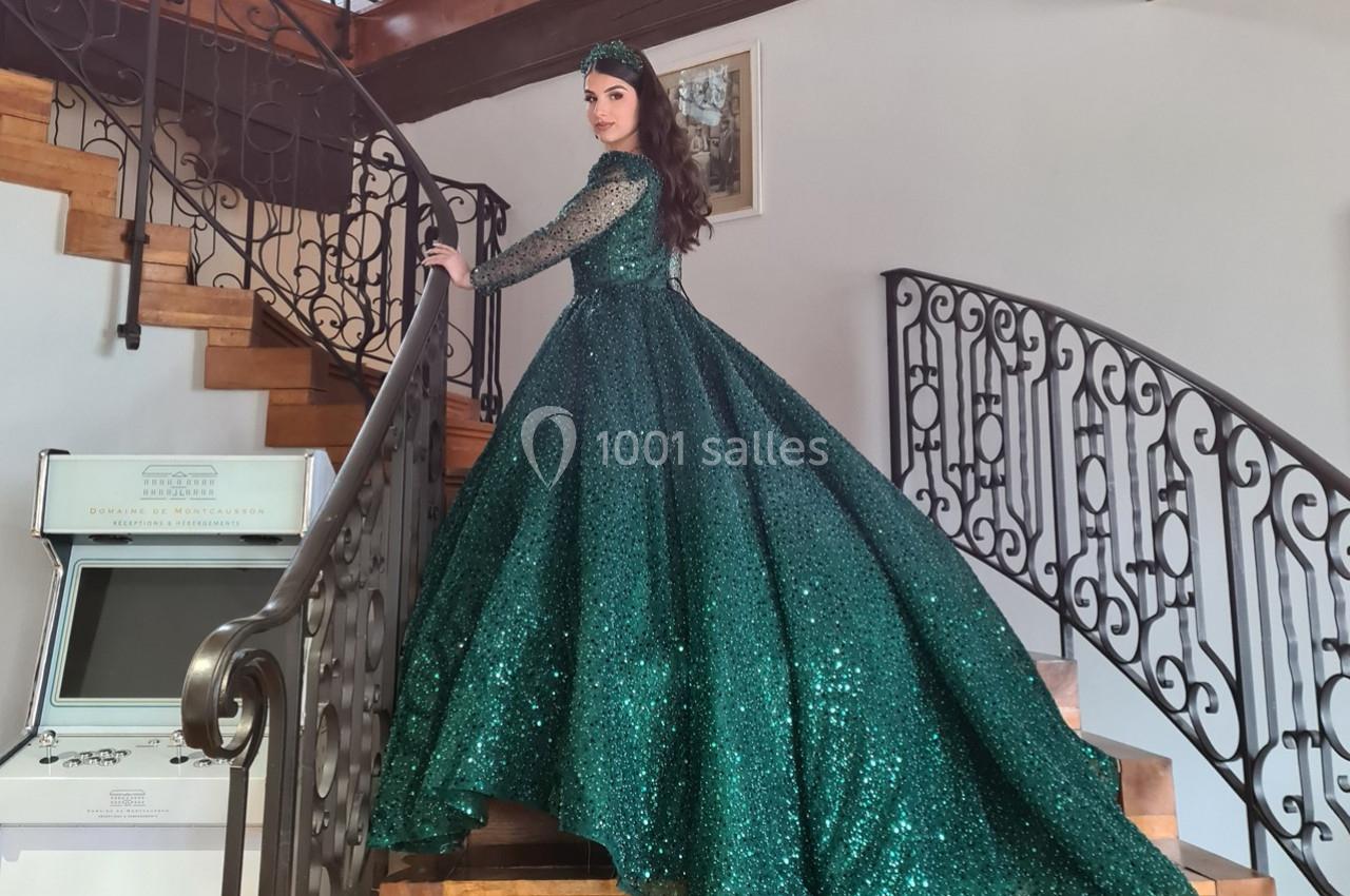 Femme en robe verte scintillante descendant un escalier en bois avec une rampe en fer forgé.