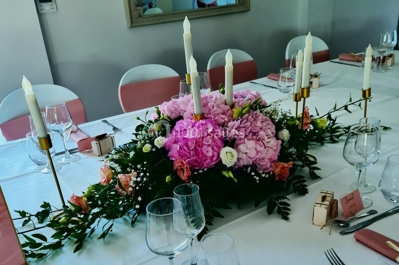 Centre de table floral avec hortensias roses, bougies blanches et vaisselle disposée sur une nappe blanche.