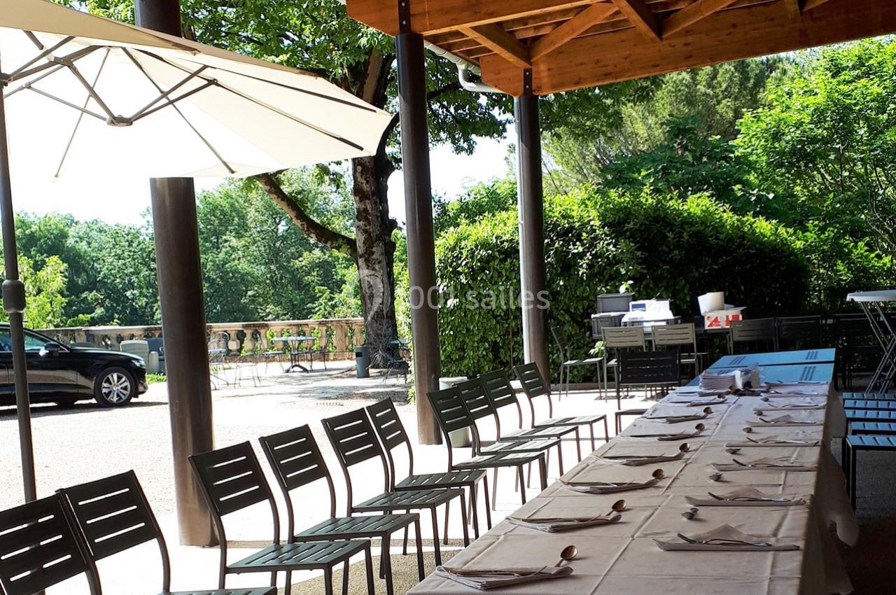 Tables dressées avec des nappes blanches et chaises alignées sous une terrasse ombragée, vue sur un jardin verdoyant.