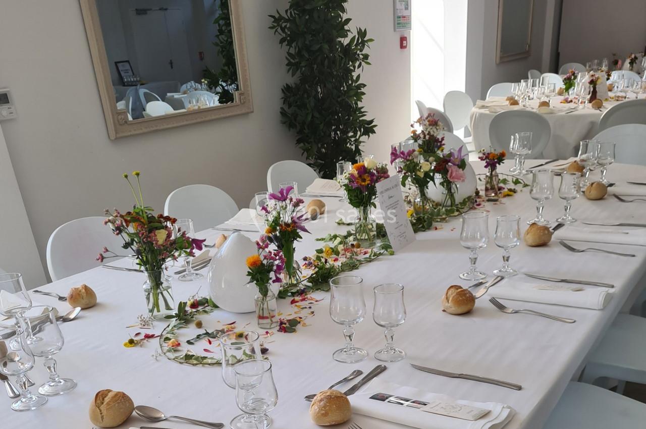 Table dressée pour un repas festif avec nappes blanches, fleurs colorées, verres, couverts et petits pains.