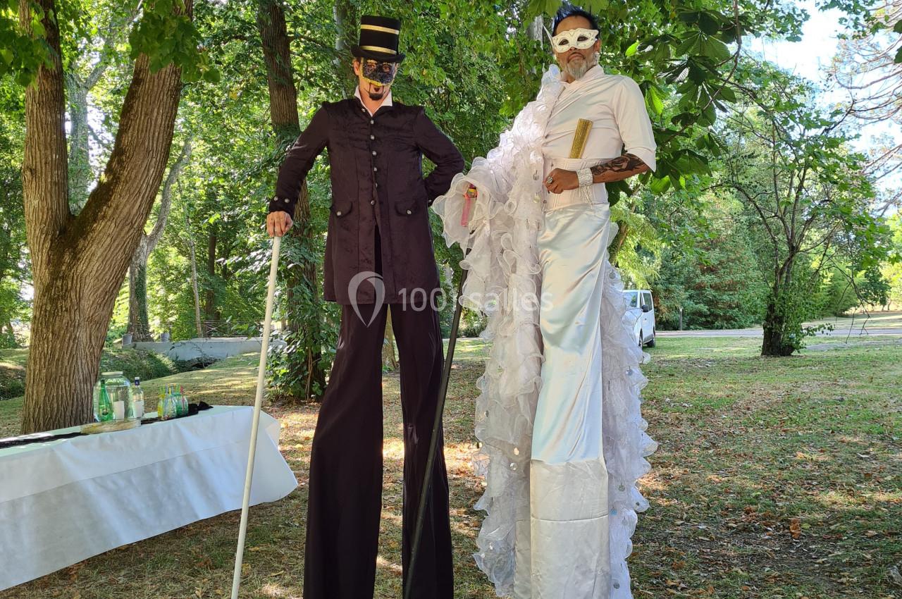 Deux personnes sur échasses, vêtues de costumes élaborés et masques, posent dans un parc verdoyant.