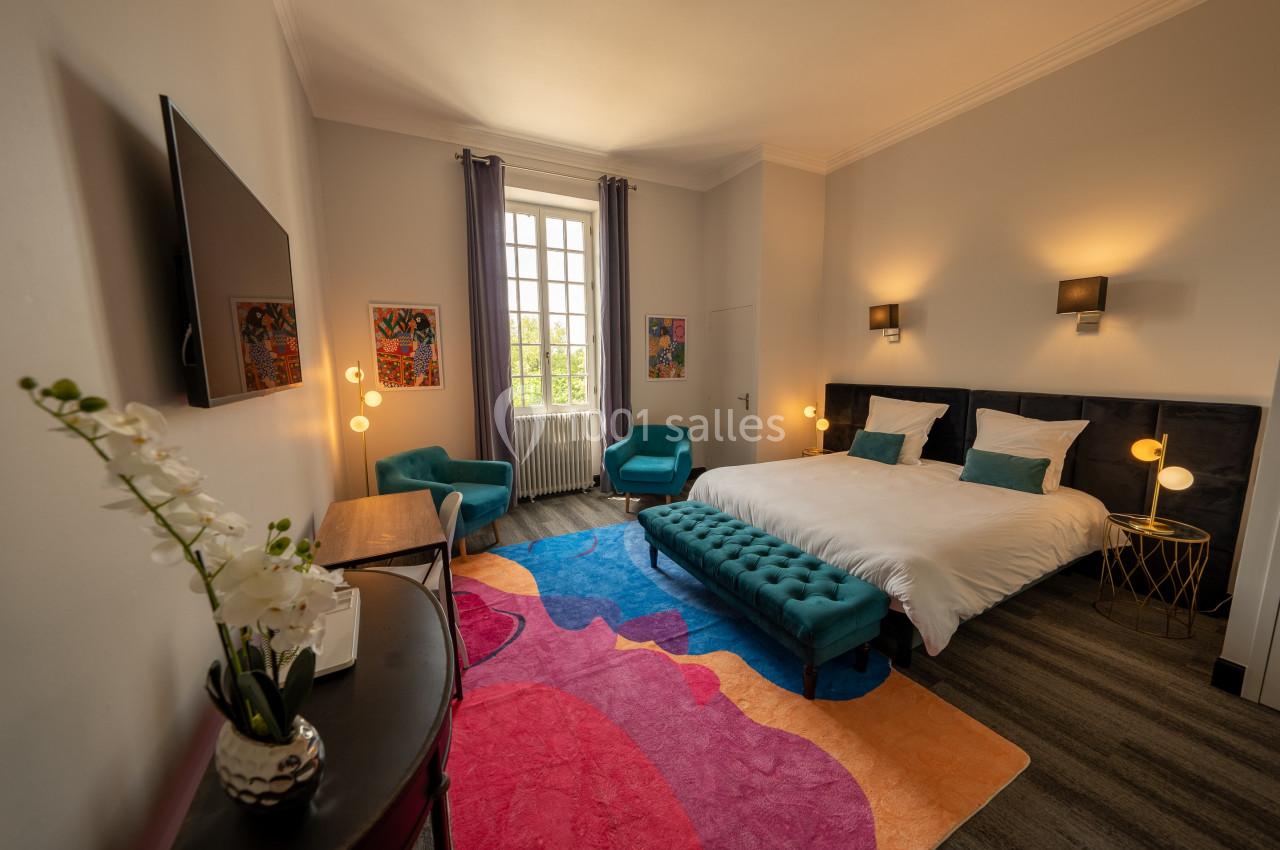 Chambre lumineuse avec lit double, fauteuils, tapis coloré, orchidée sur une table et grande fenêtre avec vue.