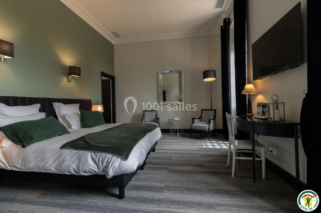 Chambre moderne avec lit double, murs verts, bureau, miroir, fauteuils et éclairage tamisé.