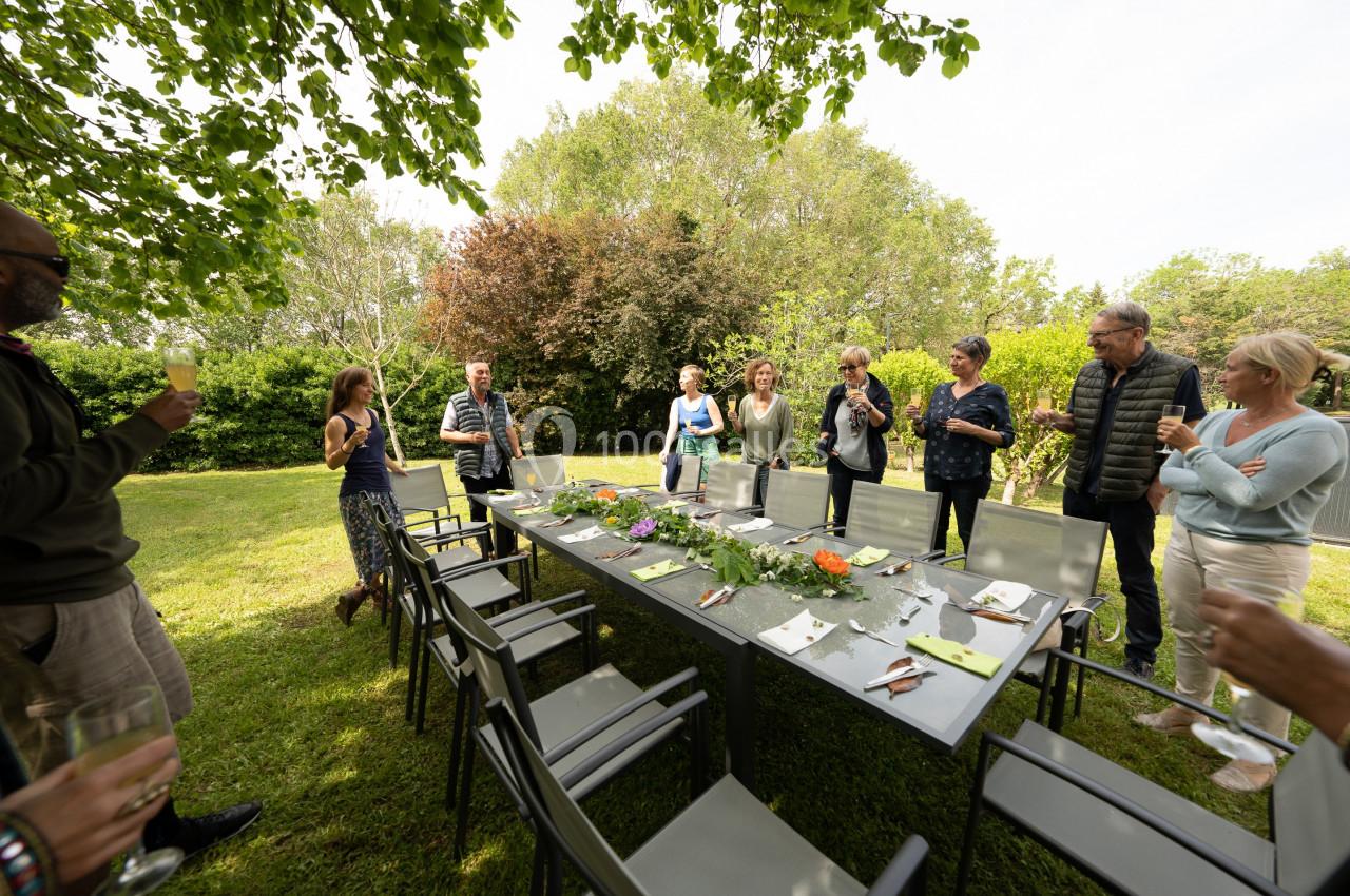 Groupe de personnes debout autour d'une table dressée dans un jardin, discutant sous des arbres par une journée ensoleillée.