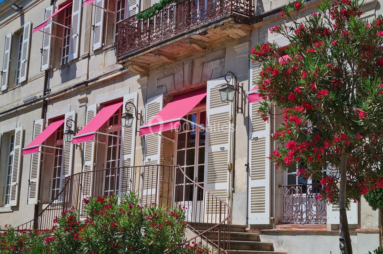 Façade d'une maison en pierre avec volets blancs, stores roses et un jardin fleuri au premier plan.