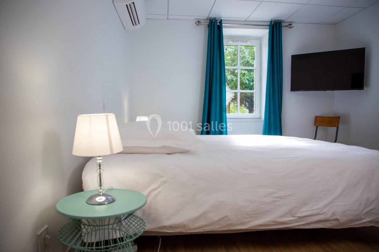 Chambre lumineuse avec lit double, table de chevet, lampe, fenêtre avec rideaux bleus et télévision fixée au mur.