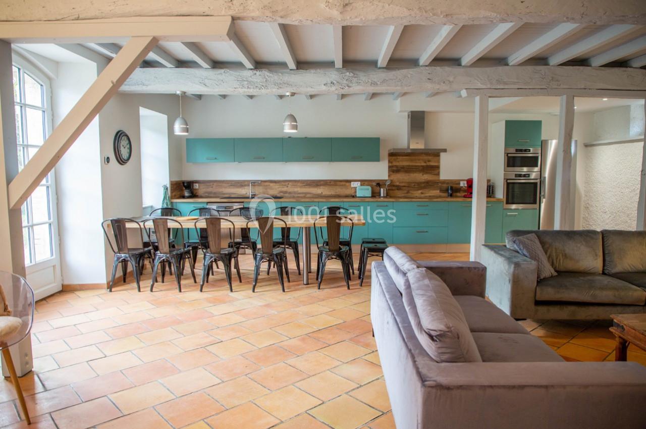 Cuisine ouverte avec meubles turquoise, grande table en bois entourée de chaises noires, et salon avec canapés gris.