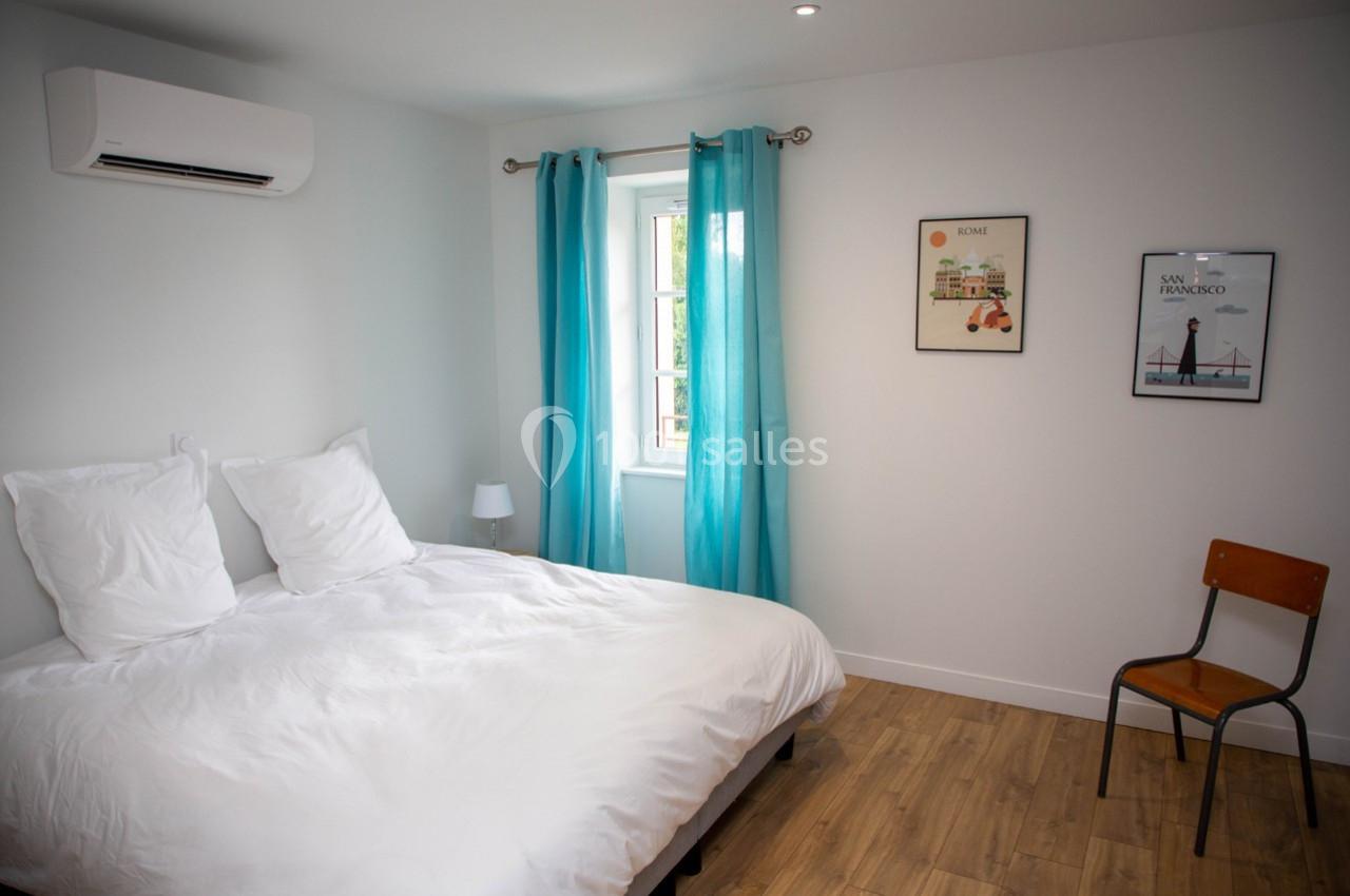 Chambre lumineuse avec lit double, rideaux bleus, chaise en bois, affiches murales et sol en parquet clair.