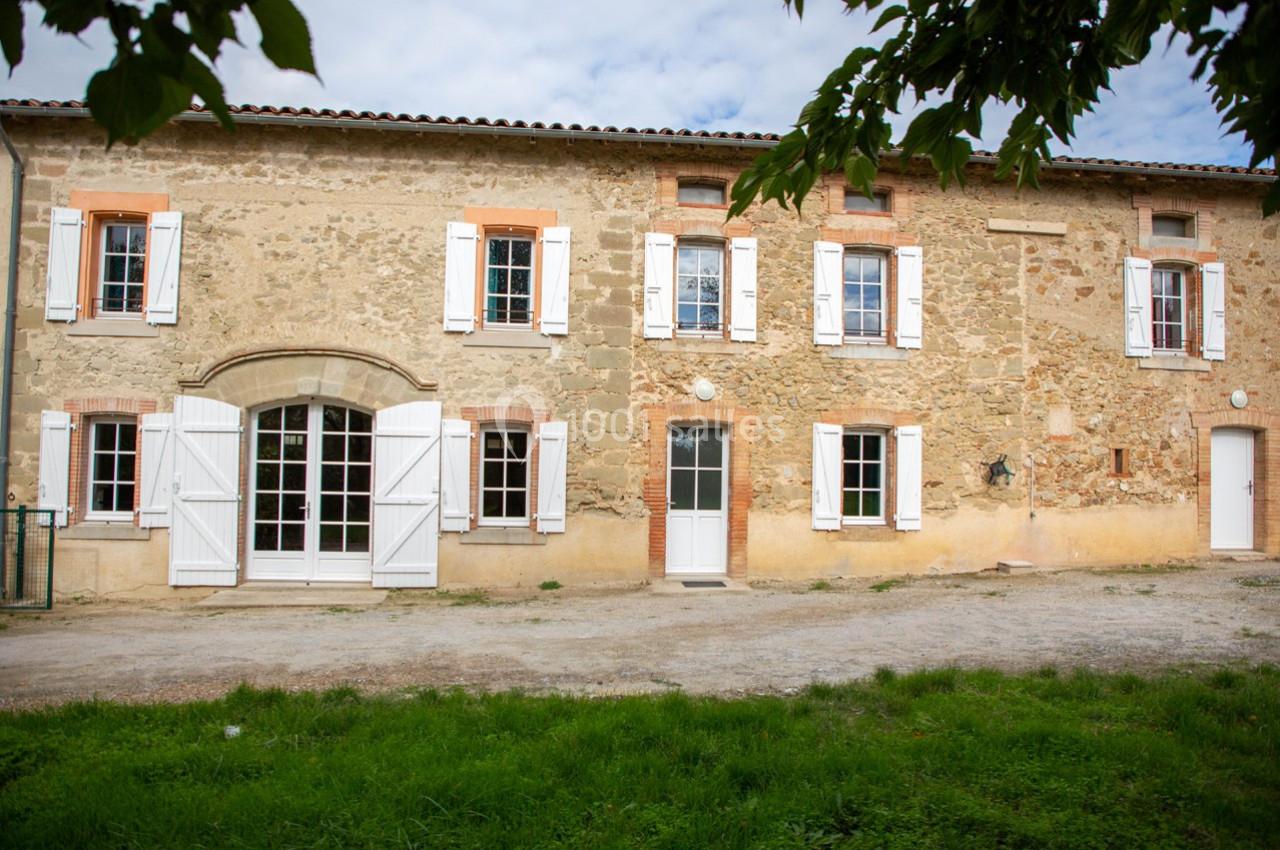 Façade d'une maison en pierre avec volets blancs, plusieurs fenêtres et une porte, entourée de verdure.