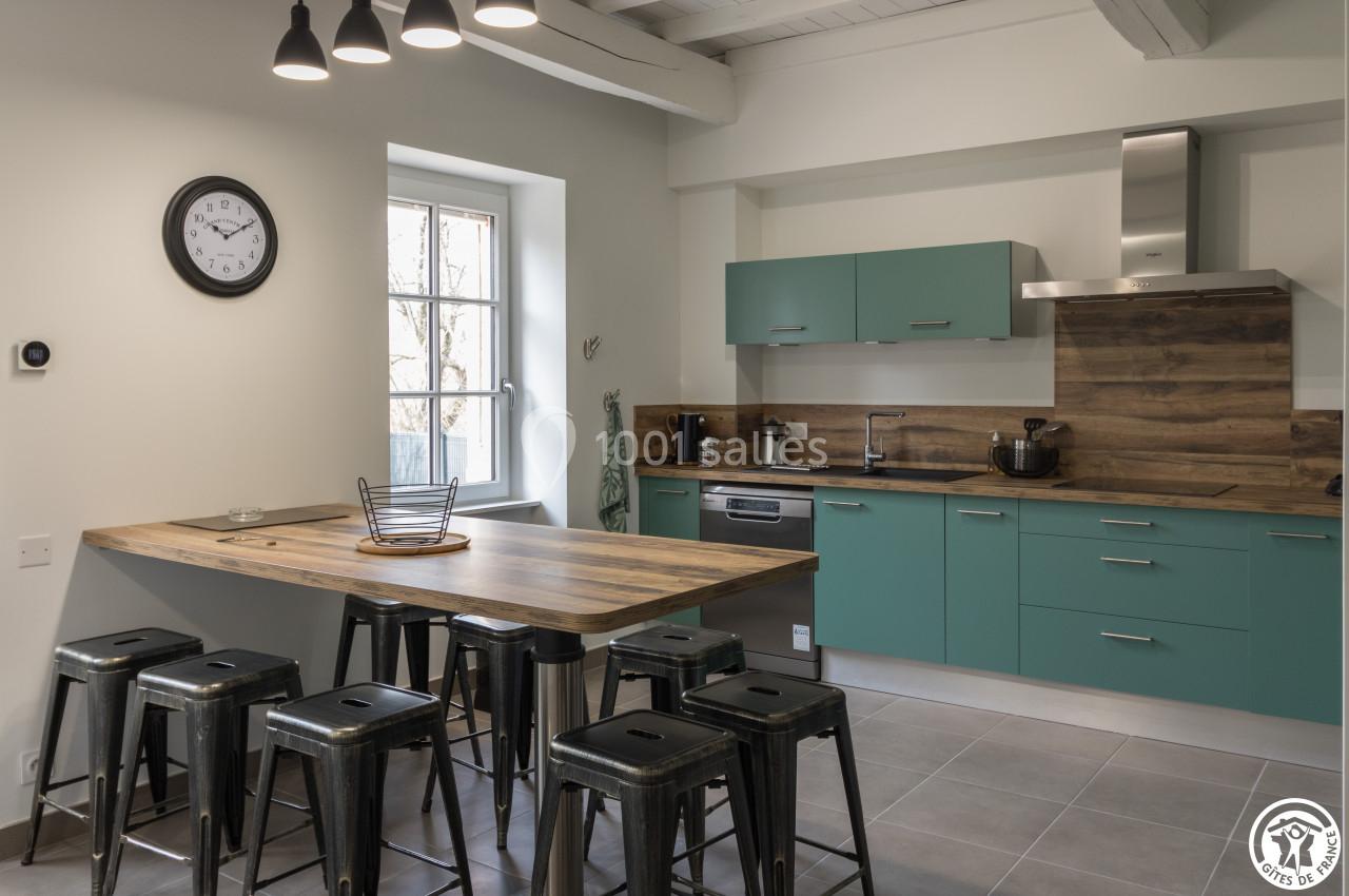 Cuisine moderne avec meubles verts, plan de travail en bois, tabourets métalliques et éclairage suspendu.