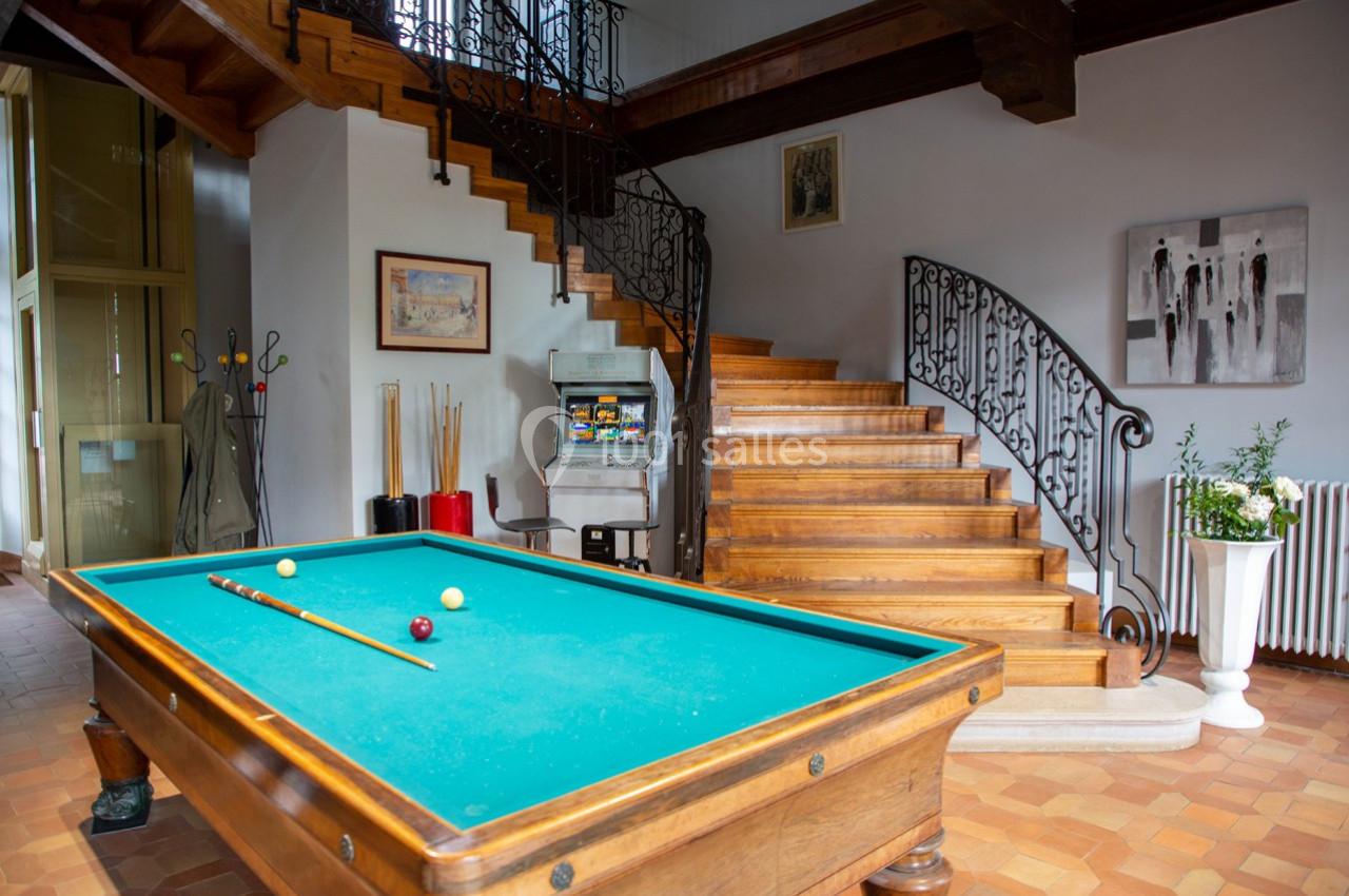 Table de billard dans une pièce lumineuse avec escalier en bois, décorations murales et sol en carrelage.