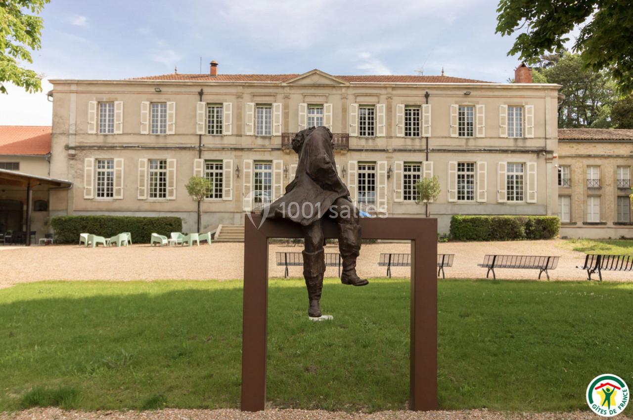 Statue d'un homme assis sur un cadre métallique devant un bâtiment ancien avec des fenêtres symétriques.