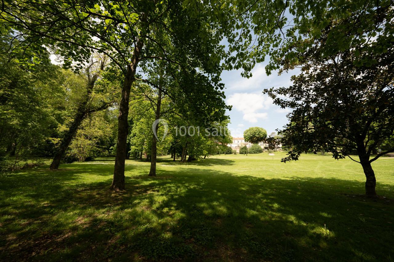 Pelouse verdoyante entourée d'arbres sous un ciel partiellement nuageux, avec un bâtiment visible au loin.