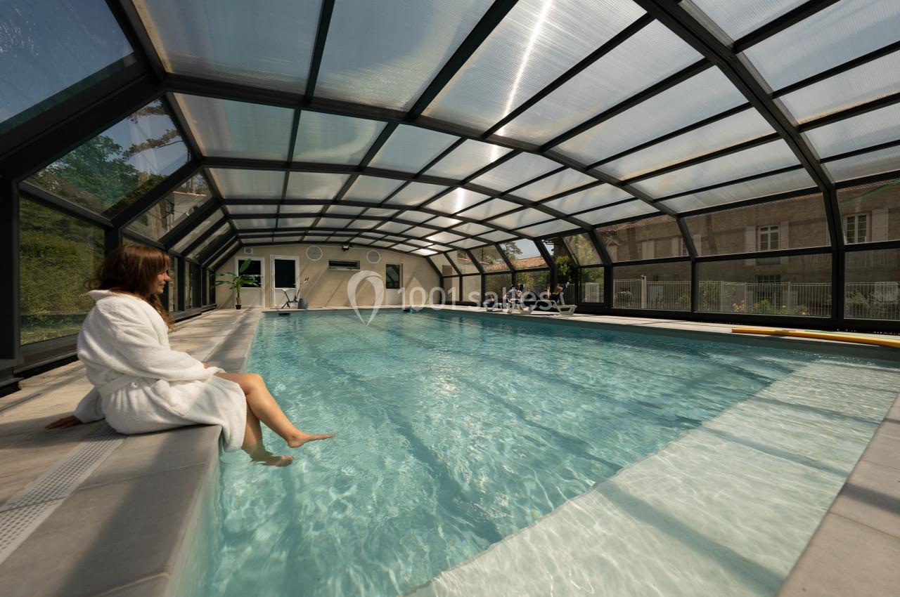 Femme en peignoir assise au bord d'une piscine couverte, avec des baigneurs en arrière-plan.