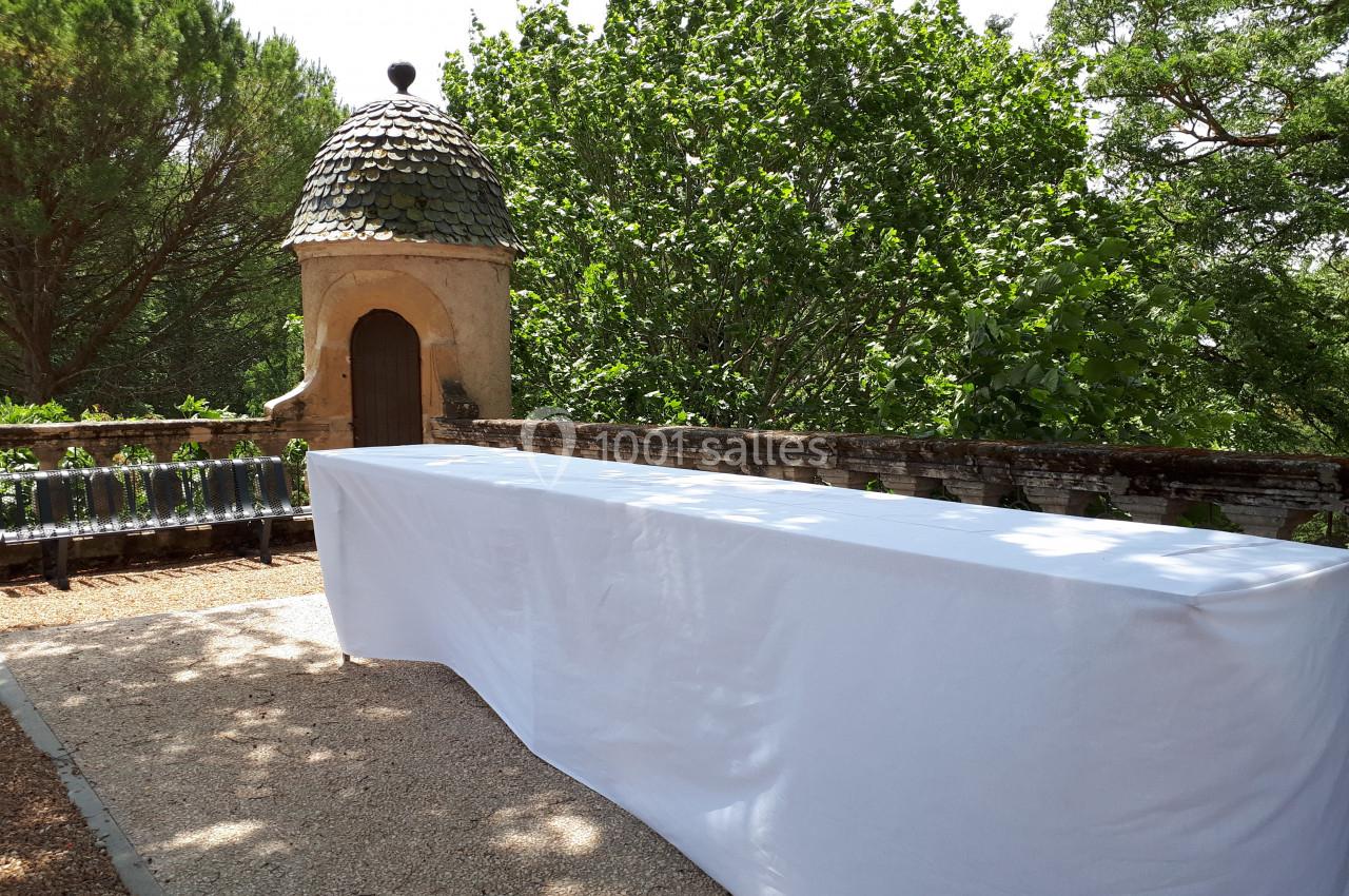 Table rectangulaire recouverte d'une nappe blanche, placée en extérieur près d'un mur en pierre et d'un jardin verdoyant.