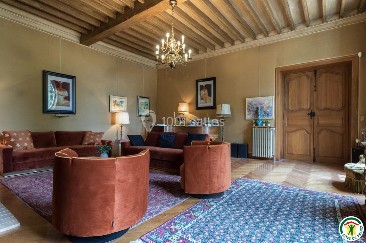 Salon chaleureux avec poutres apparentes, mobilier en velours rouge, tapis colorés et éclairage par un lustre central.