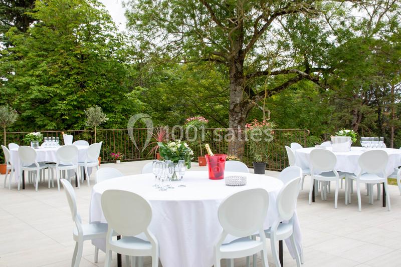 Terrasse aménagée avec des tables rondes dressées, nappes blanches, chaises modernes et végétation en arrière-plan.