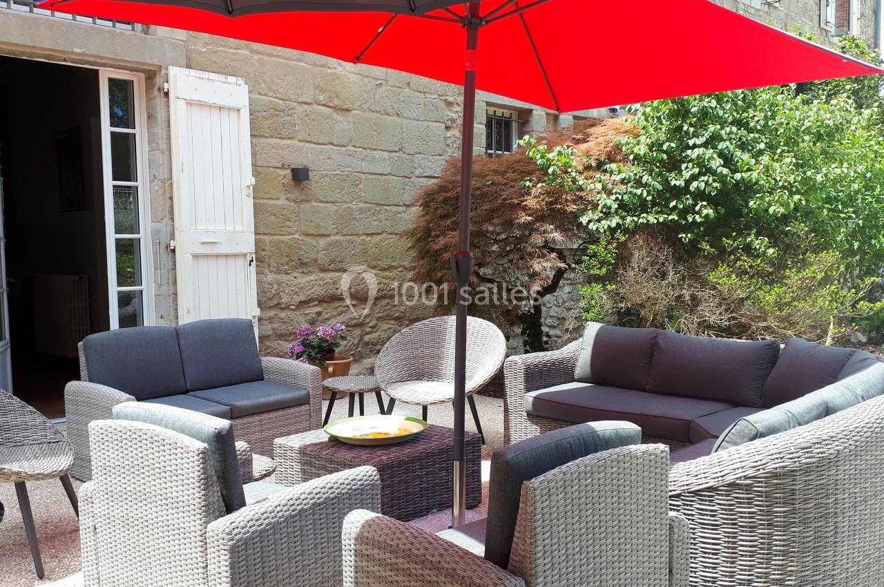 Salon de jardin en rotin avec coussins gris, disposé sur une terrasse ensoleillée, sous un grand parasol rouge.