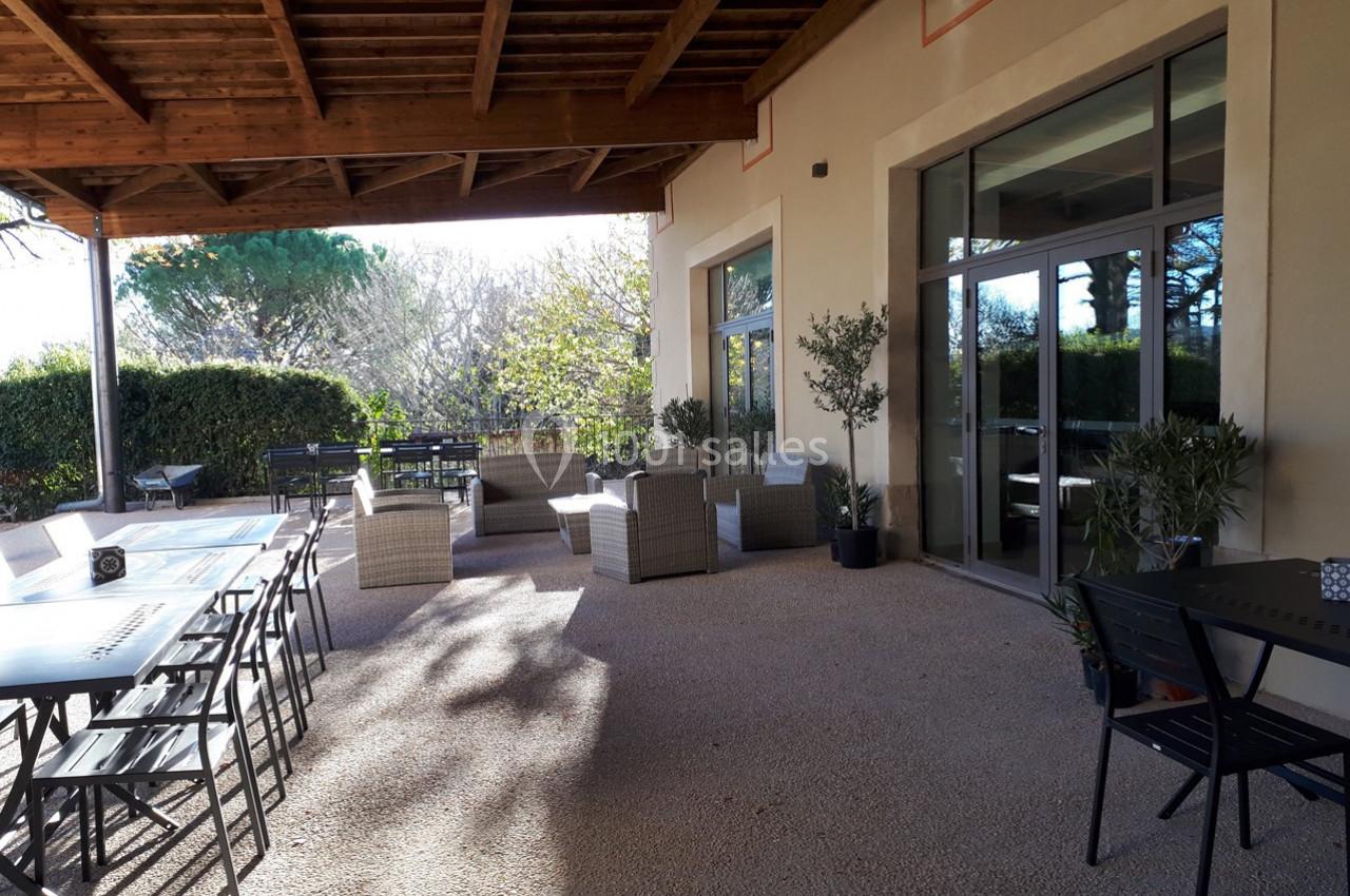 Terrasse extérieure avec tables, chaises et fauteuils sous une pergola, entourée de verdure et baignée de lumière naturelle.