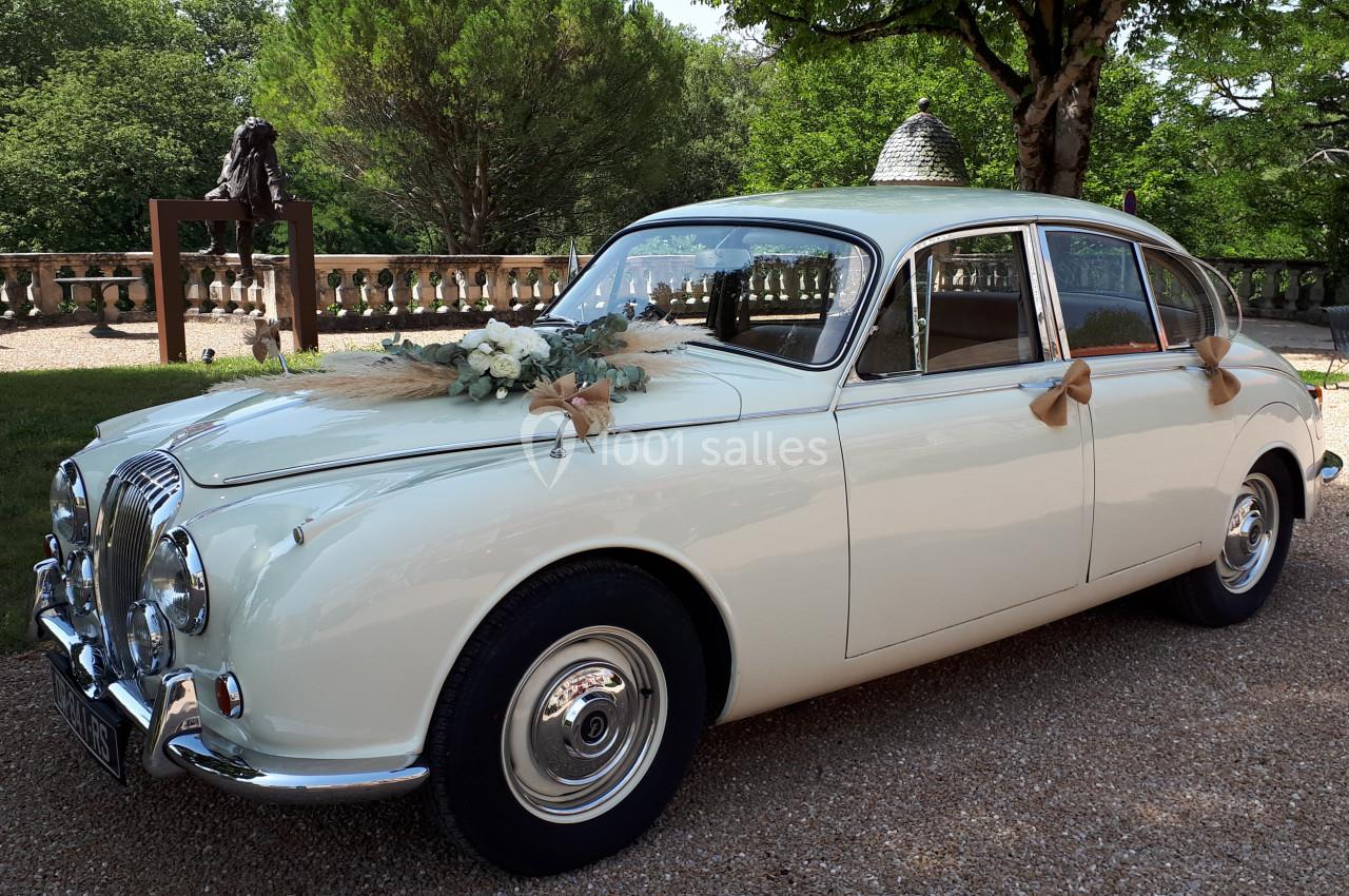 Voiture ancienne blanche décorée de fleurs et de rubans, stationnée dans une allée gravillonnée devant un jardin.