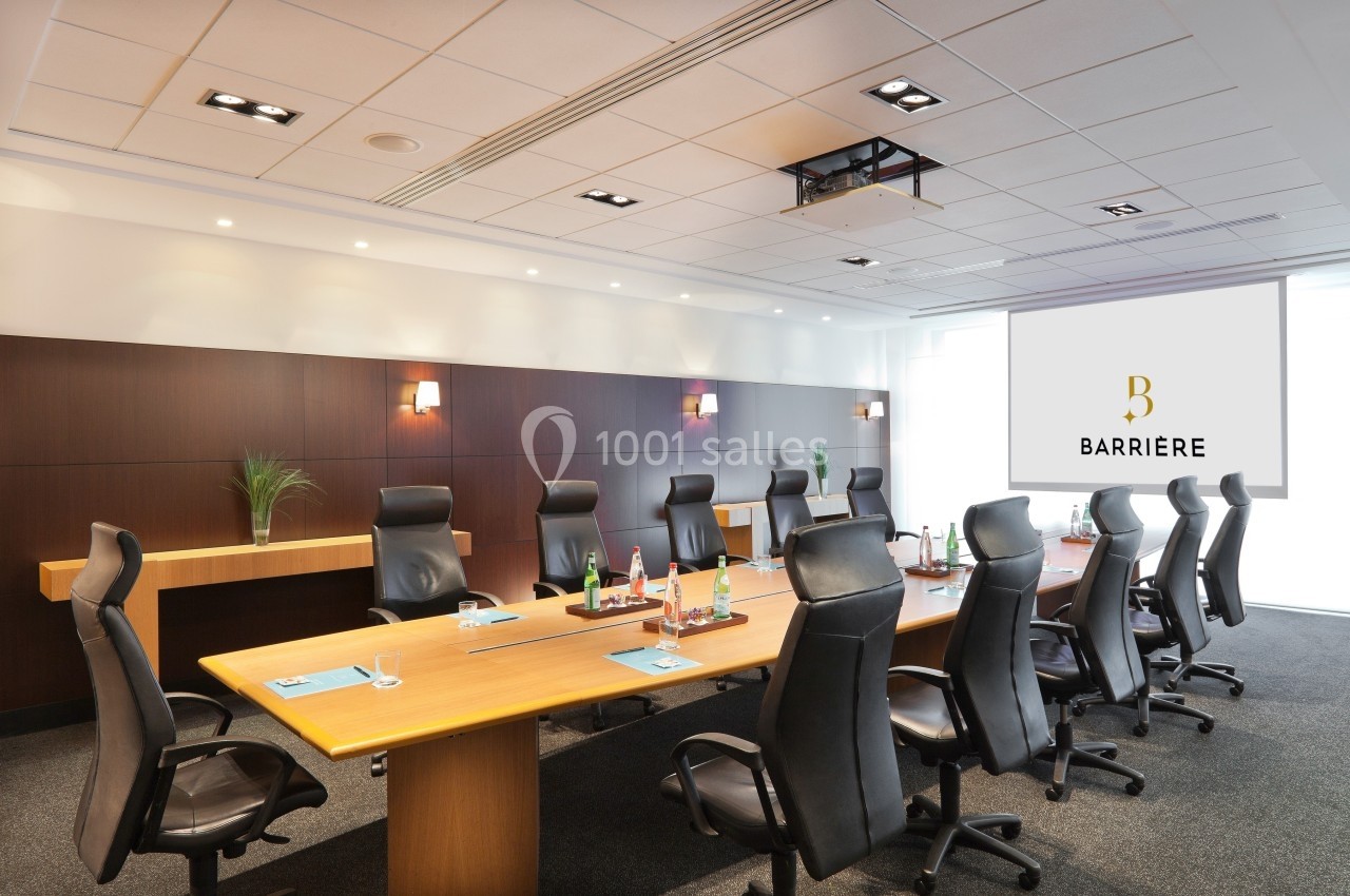 Salle de réunion moderne avec une grande table en bois, chaises noires, écran mural et éclairage intégré.