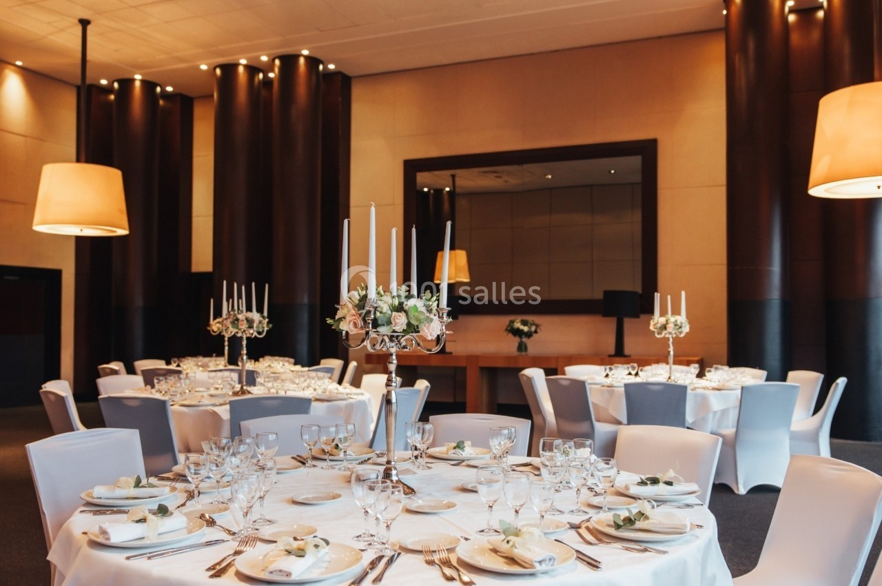 Salle de réception élégante avec tables rondes dressées, chandeliers et décoration florale, éclairage chaleureux.