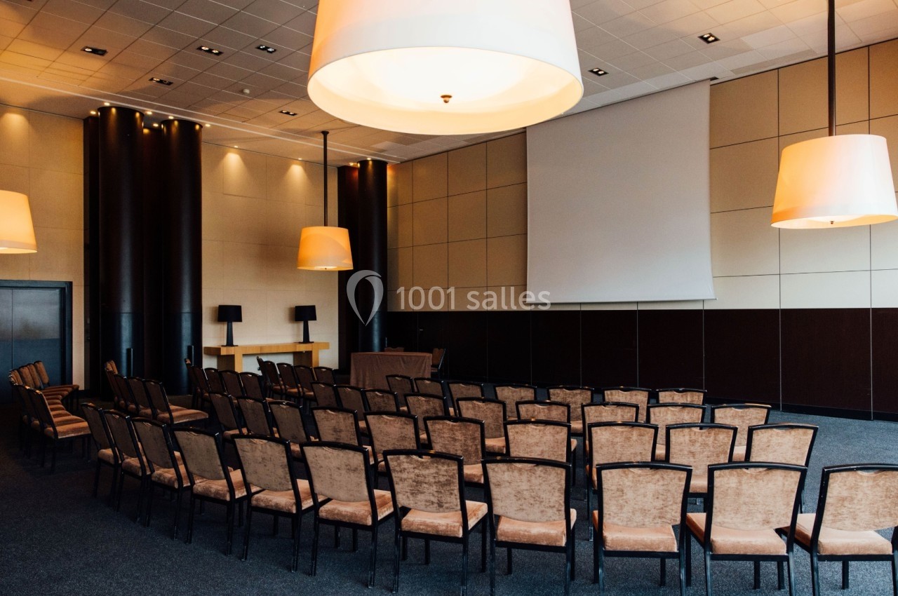 Salle de conférence vide avec des rangées de chaises alignées face à un écran de projection et un pupitre.