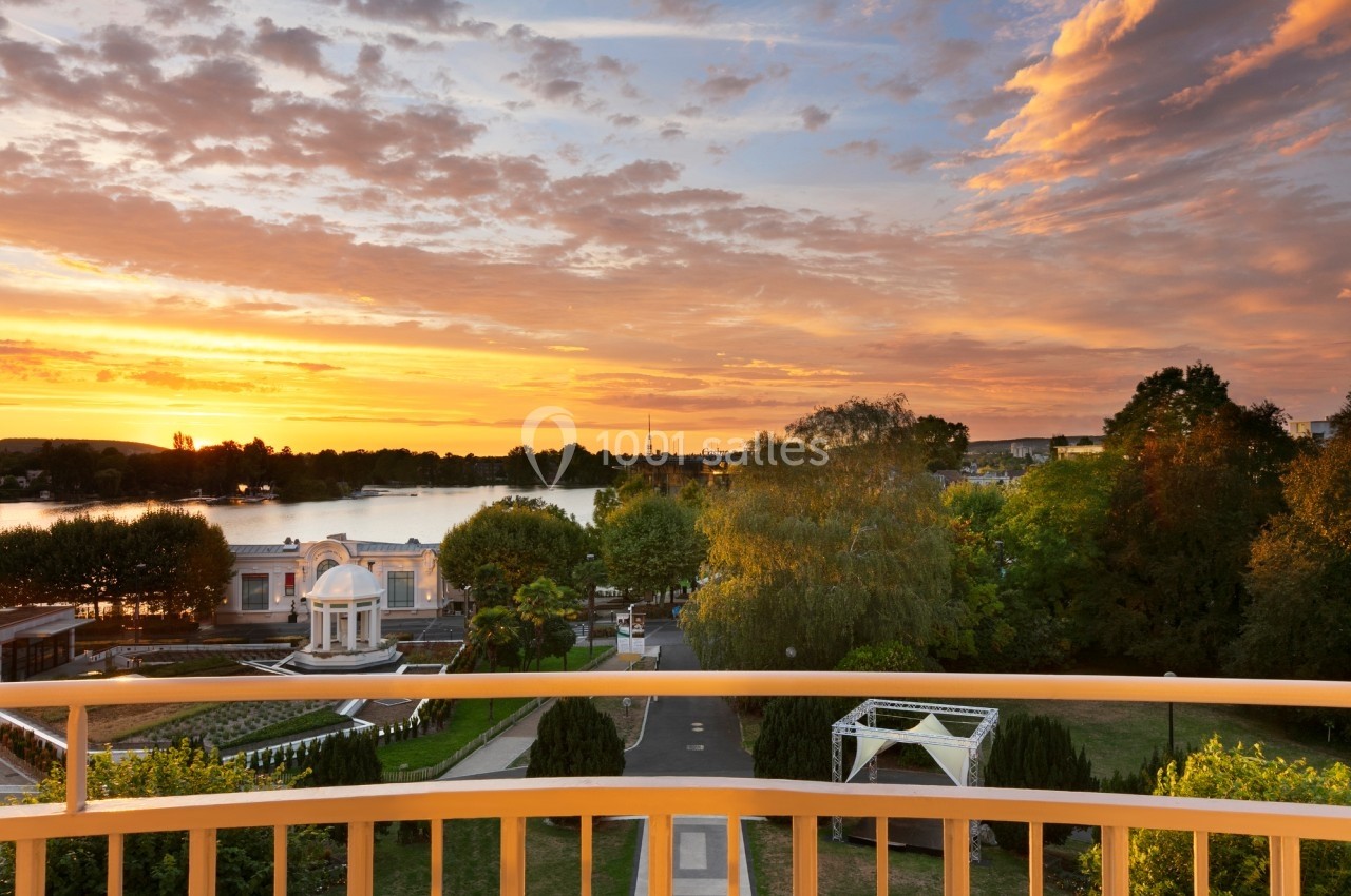 Vue d'un parc avec un pavillon, des arbres et un lac au coucher du soleil, depuis une balustrade jaune.