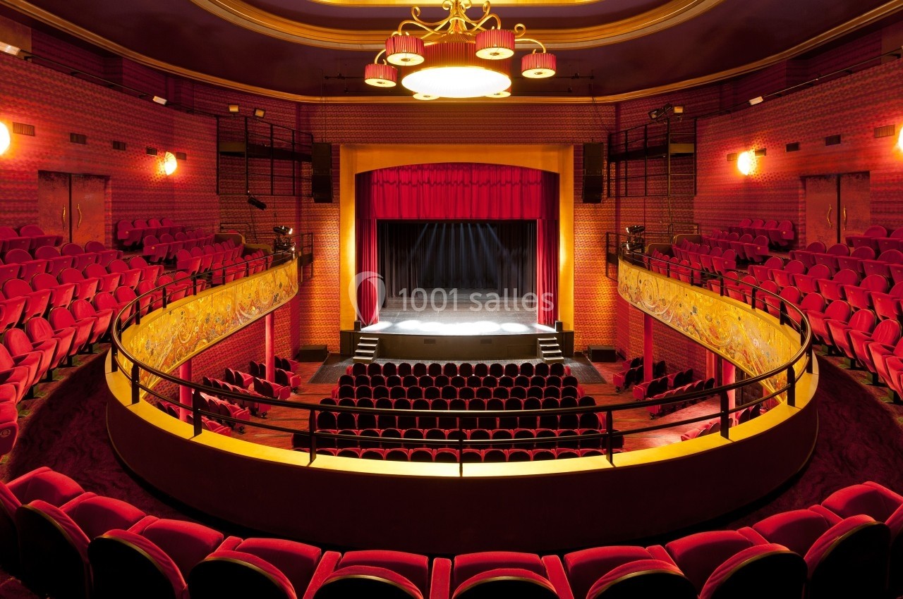 Salle de théâtre vide avec sièges rouges, scène éclairée par des projecteurs et balcons ornés de motifs dorés.