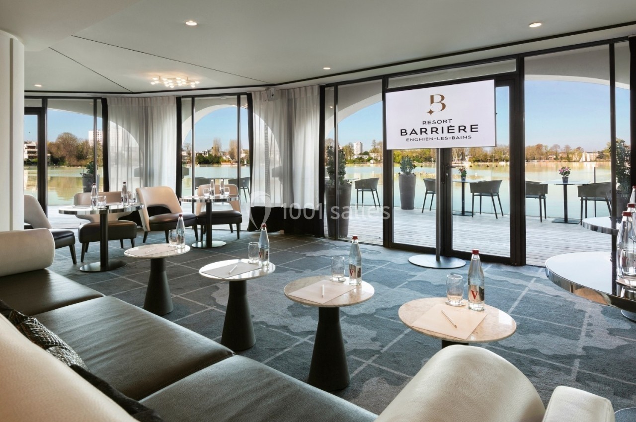 Salle lumineuse avec baies vitrées donnant sur une terrasse et un plan d'eau, aménagée avec tables, fauteuils et bouteilles…