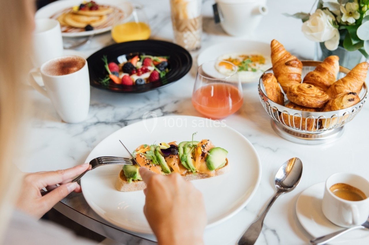 Une personne déguste une tartine garnie d'avocat et saumon, entourée de viennoiseries, fruits et boissons sur une table.