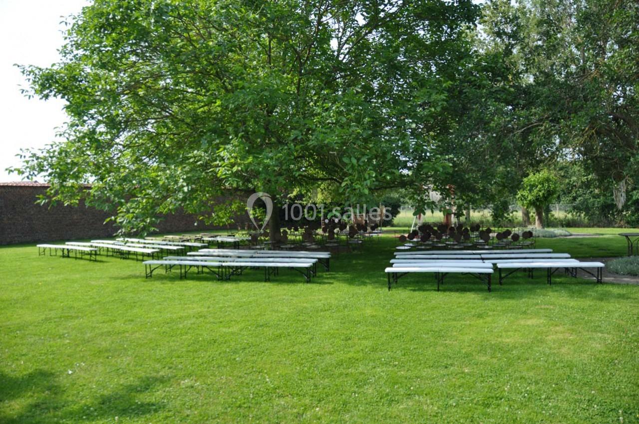Des tables et bancs installés sous des arbres sur une pelouse, dans un espace extérieur calme et verdoyant.