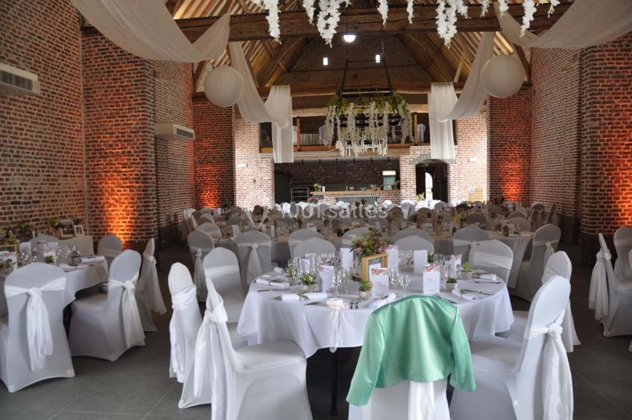 Salle de réception avec tables décorées, nappes blanches, chaises couvertes et guirlandes suspendues sous un plafond en bois.